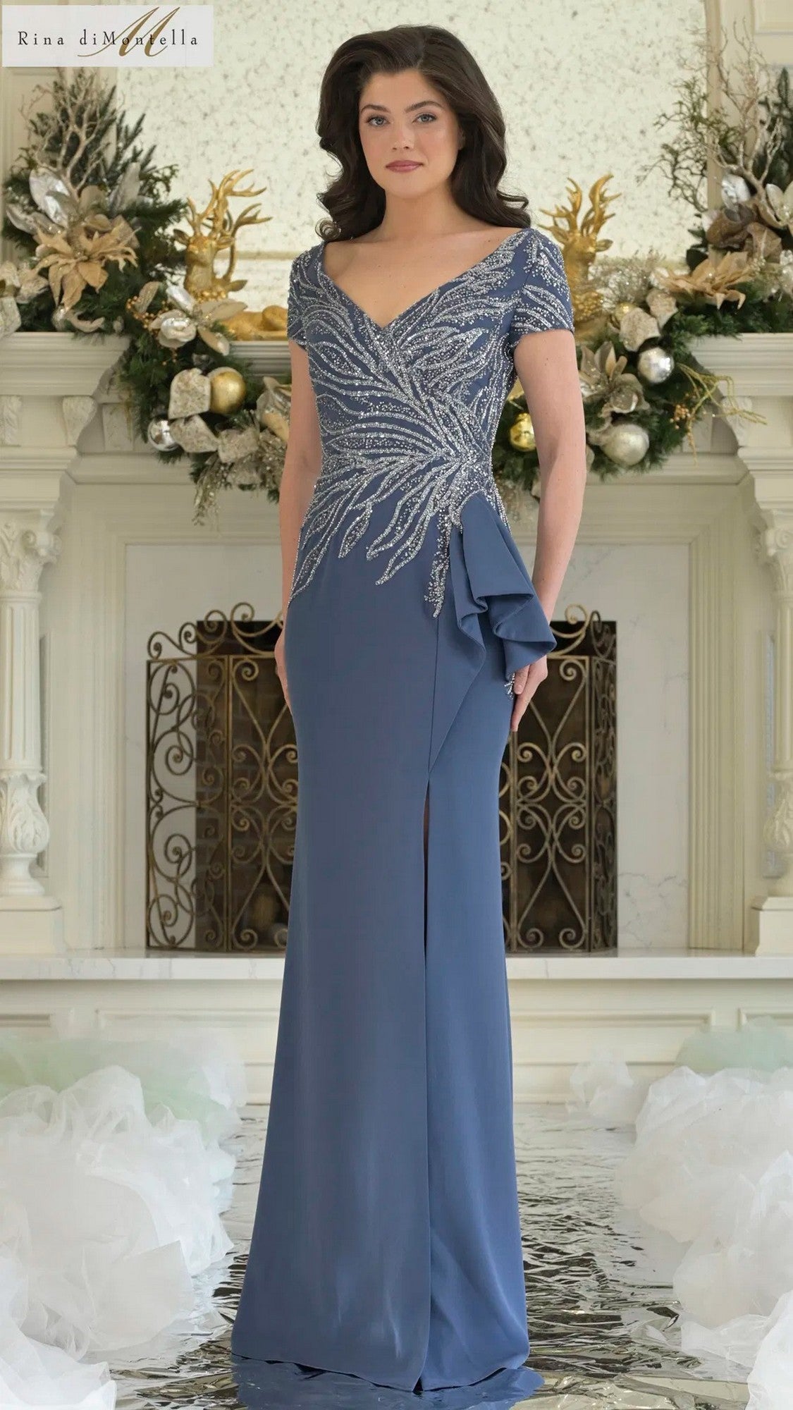 A woman in the Long Formal Dress: Rina Di Montella RD3323 in Deep Royal, featuring a fitted beaded bodice and silver embellishments, stands indoors before a fireplace with white and gold floral arrangements.